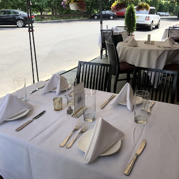 Outdoor dining tables set with white tablecloths, napkins, and silverware, with cars in the background.