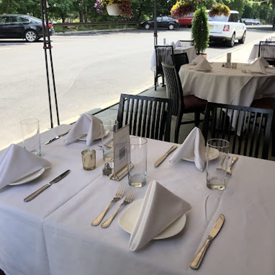 Outdoor dining tables set with white tablecloths, napkins, and silverware, with cars in the background.