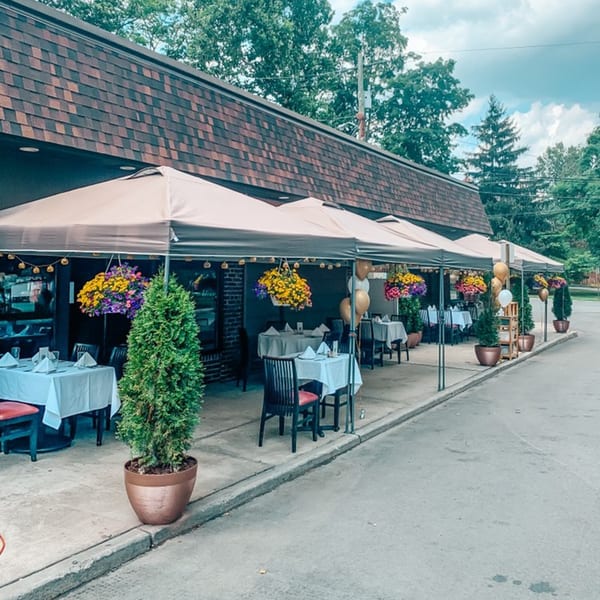 Outdoor patio dining at Cork & Crust Italian Kitchen with tables under umbrellas, hanging flowers, and potted plants.