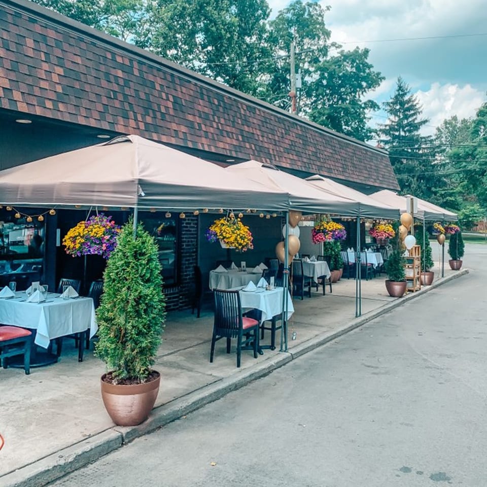 Outdoor patio dining at Cork & Crust Italian Kitchen with tables under umbrellas, hanging flowers, and potted plants.