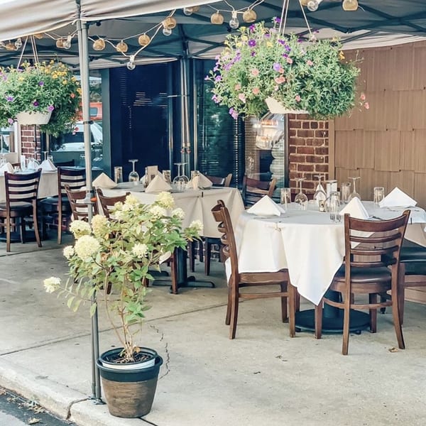 Outdoor patio dining at Cork & Crust Italian Kitchen with set tables, hanging flower baskets, and string lights.