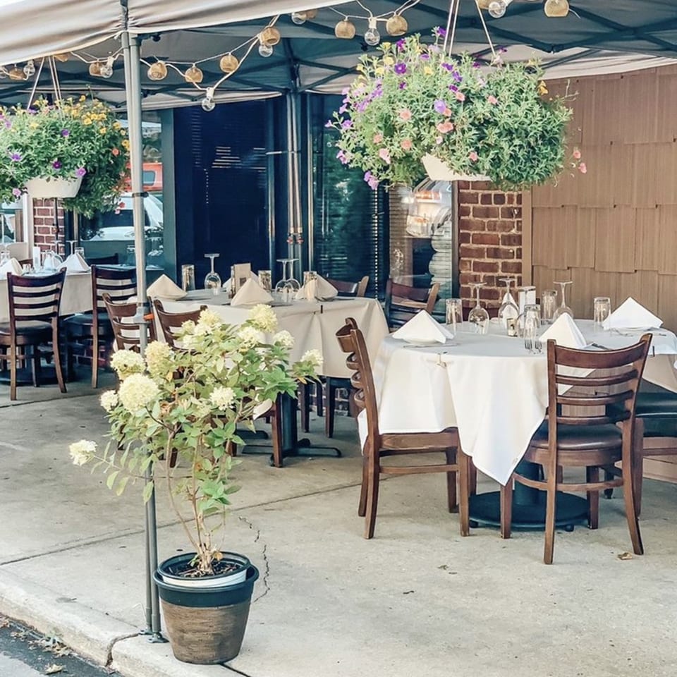 Outdoor patio dining at Cork & Crust Italian Kitchen with set tables, hanging flower baskets, and string lights.